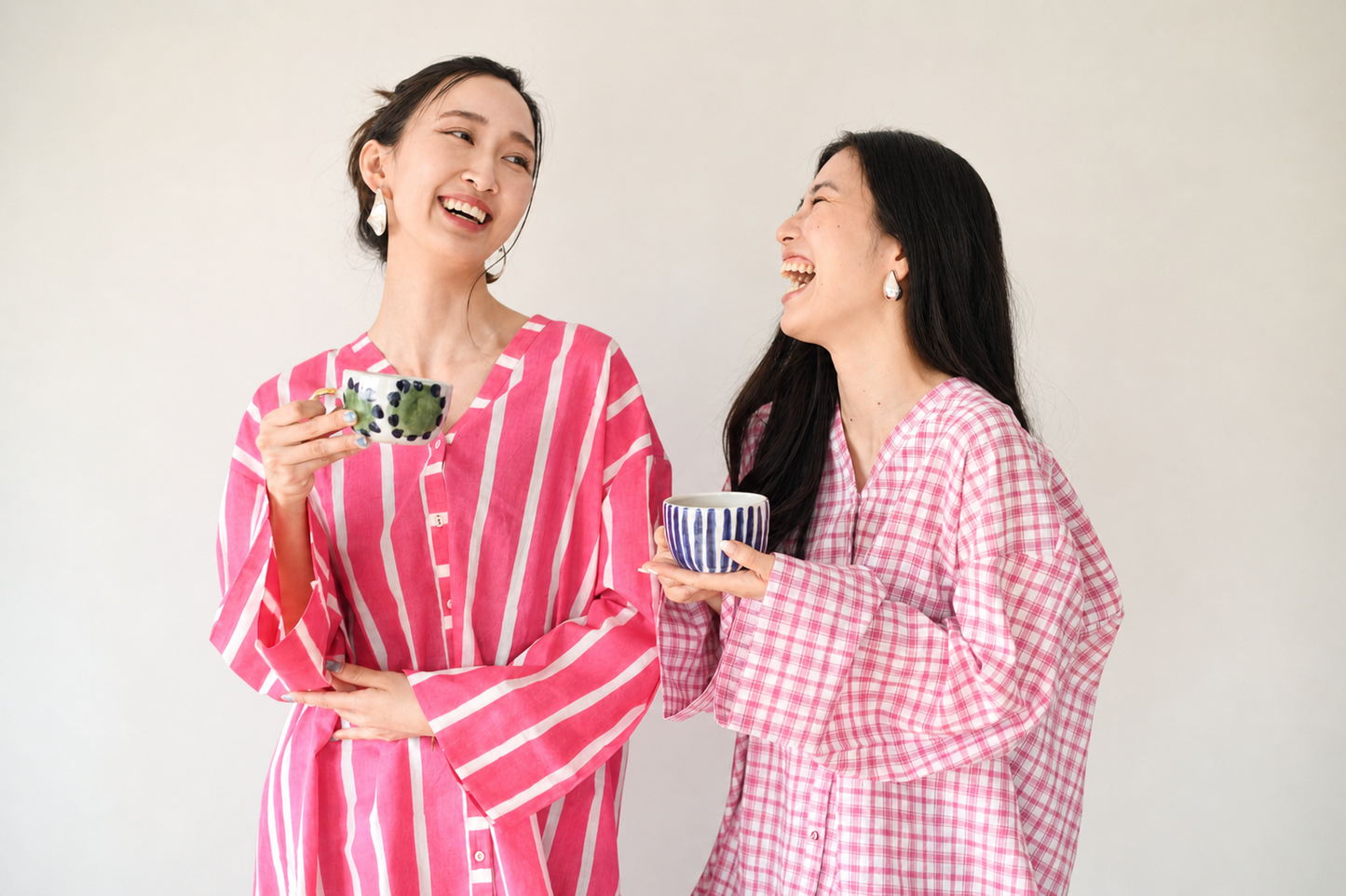 Two women in pink and white striped and checkered shirts holding mugs, laughing against a plain background.