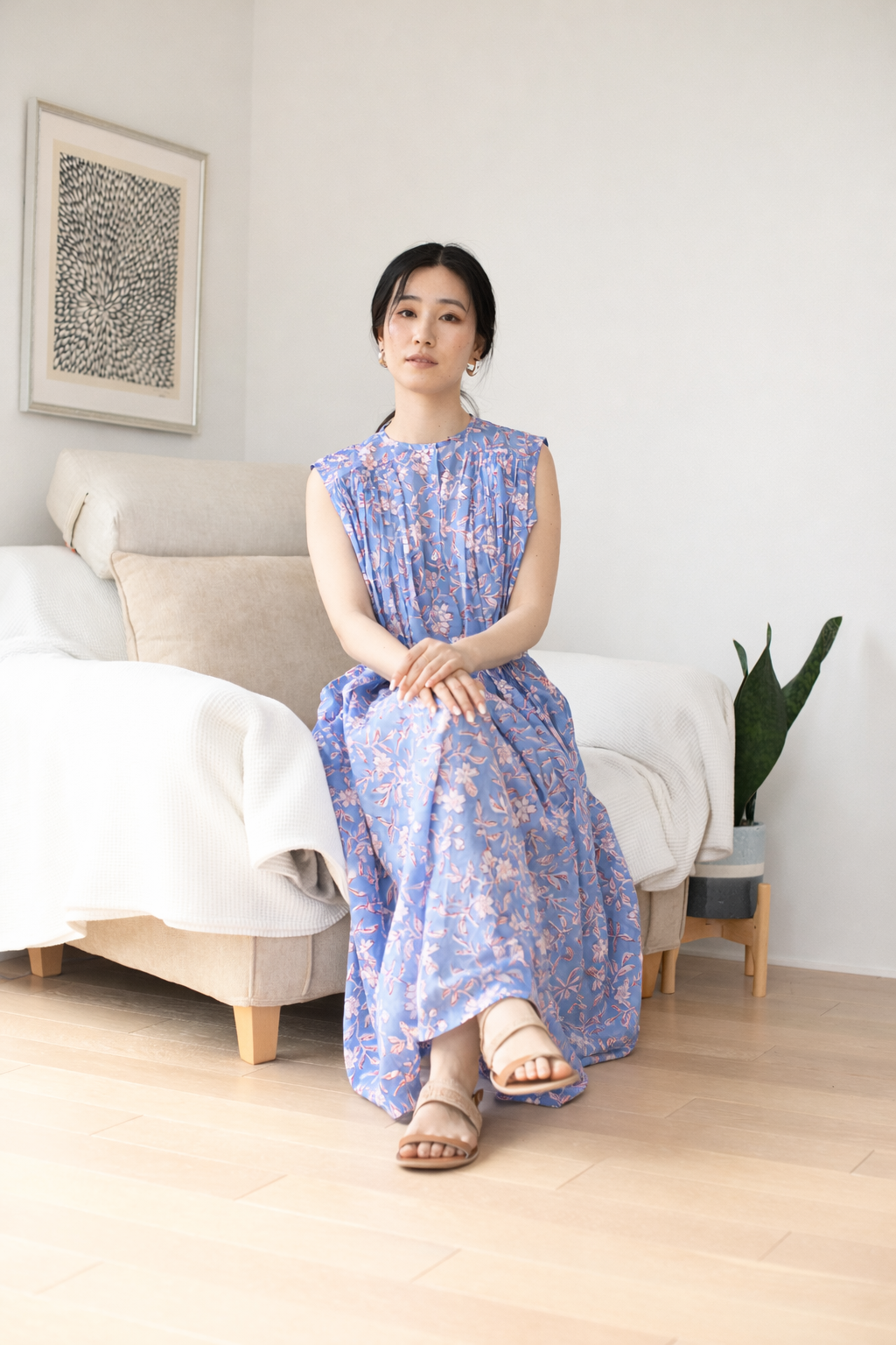 Woman in a blue and pink floral dress sitting on a couch in a room with a white wall and framed artwork.