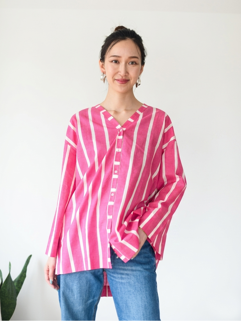 A woman wearing a pink and white vertically striped button-up shirt with blue jeans, standing against a plain white wall.