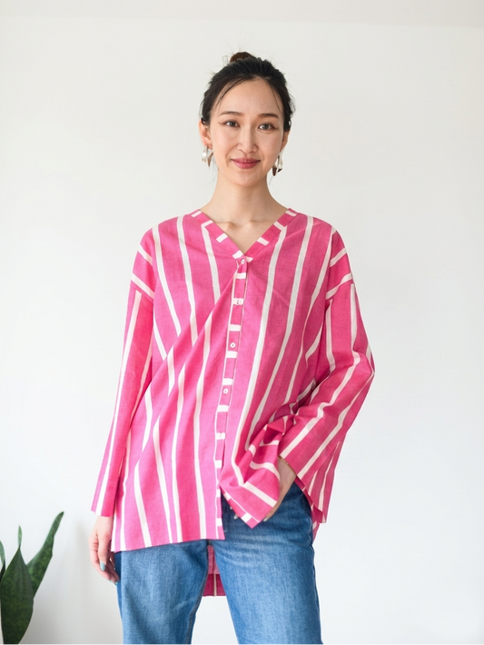 A woman wearing a pink and white vertically striped button-up shirt with blue jeans, standing against a plain white wall.
