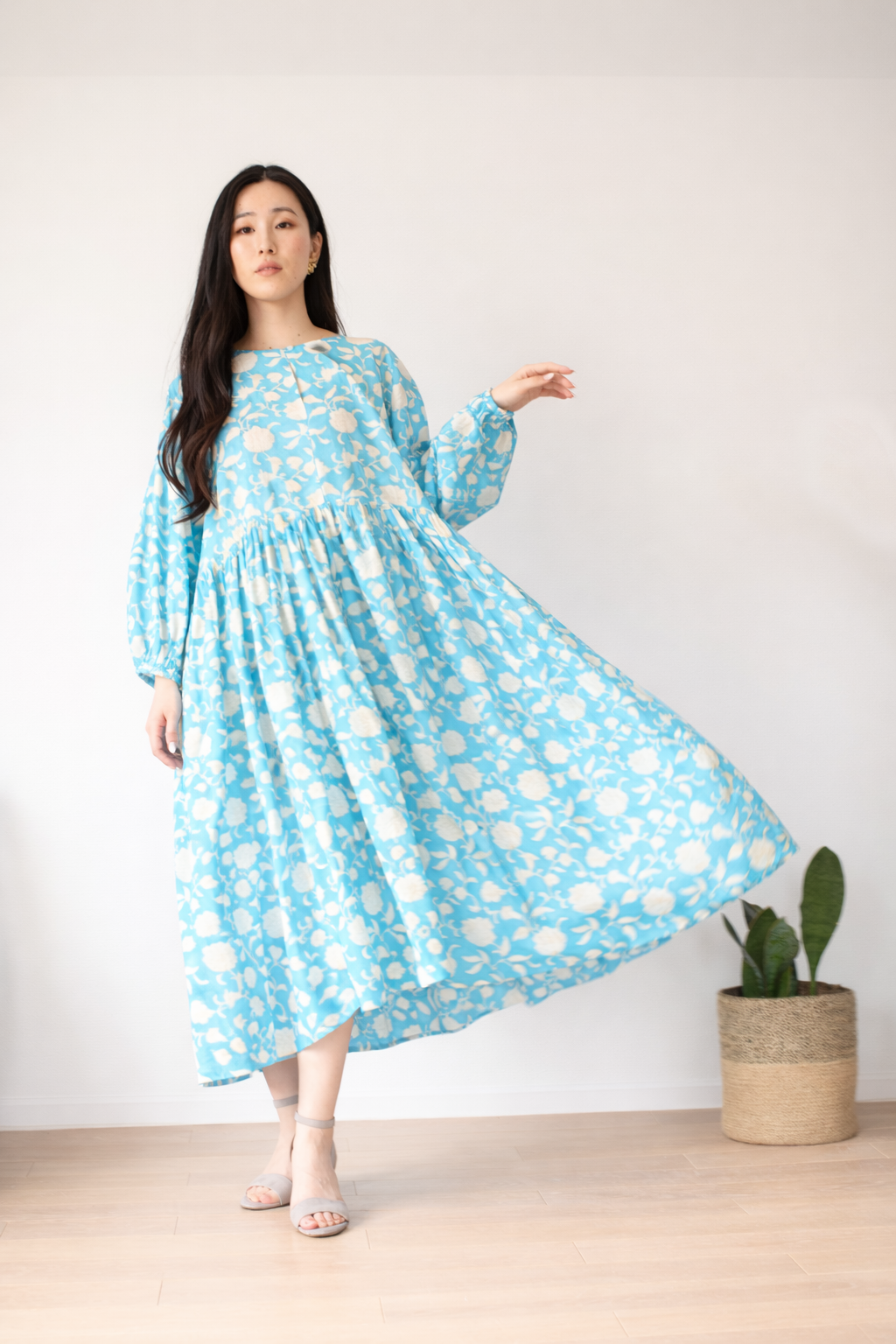 Woman wearing a turquoise blue flare dress with white floral patterns, standing indoors with a plant in the background.