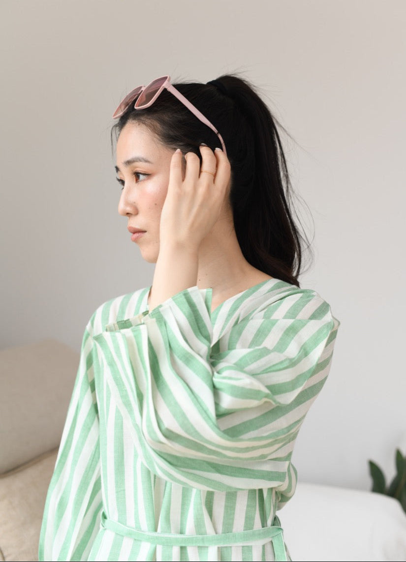 Woman wearing a green and white striped dress with sunglasses on her head, sitting against a neutral background.