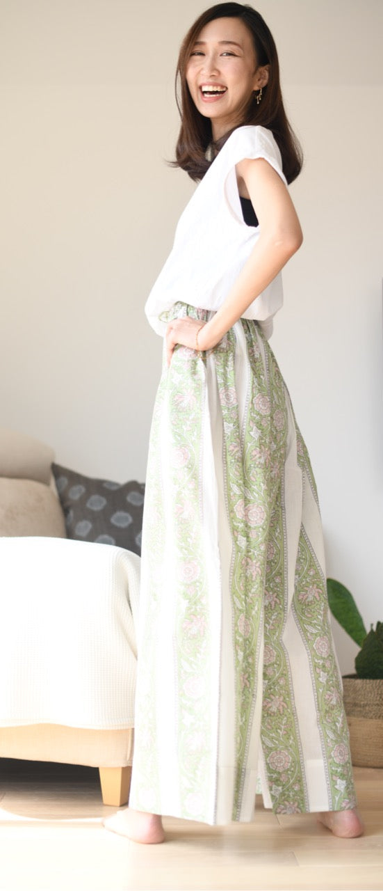 Woman wearing a white top and green patterned flare pants standing in a room with a couch and plant.